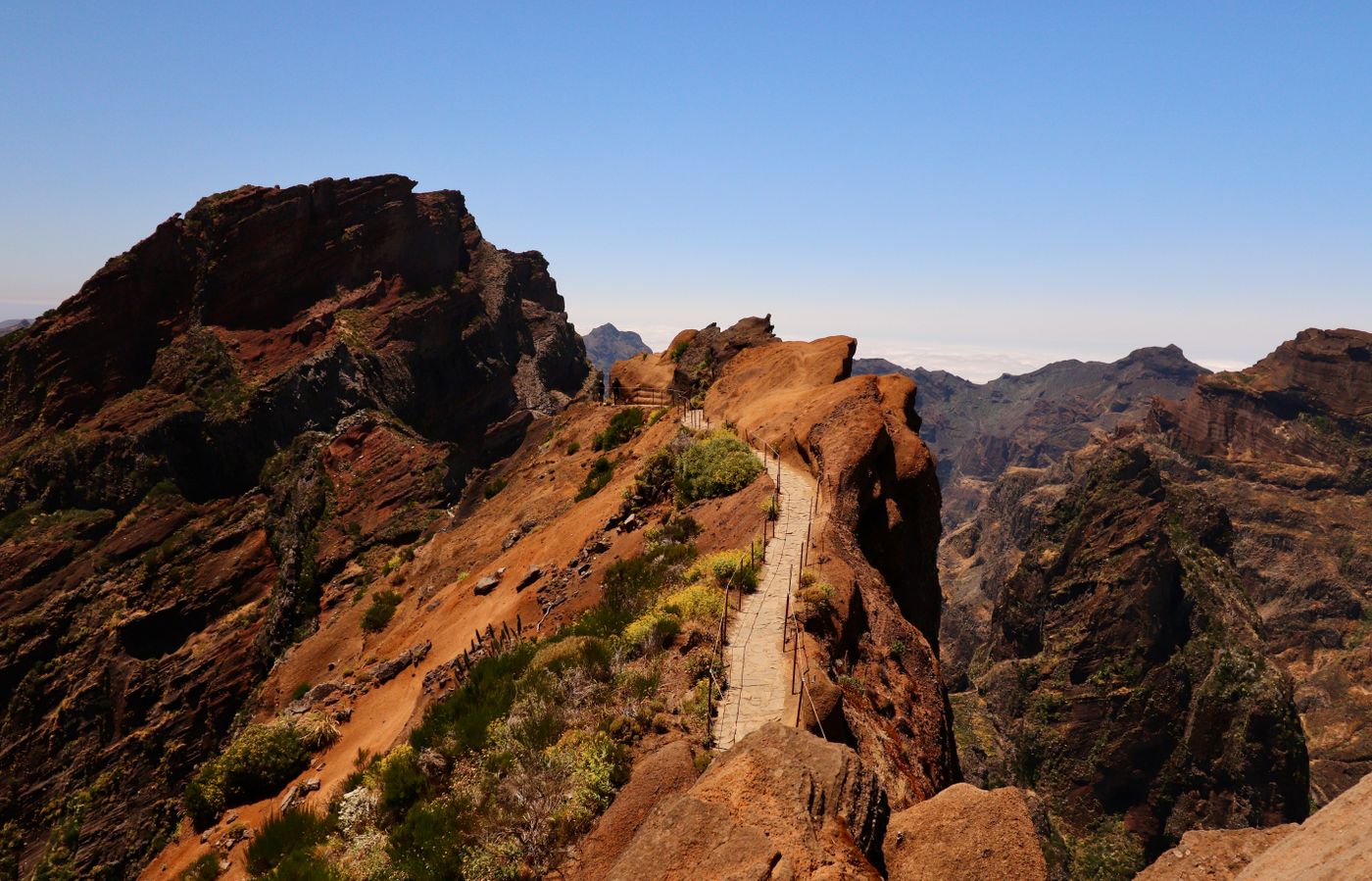A staircase of steps cut into a reddish volcanic ridge on the Pico do Arieiro to Pico Ruivo trail, dropping along a knife-edge spine toward distant valleys